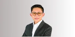 Seseorang mengenakan jas gelap dan kemeja putih, berdiri dengan pose percaya diri di latar belakang berwarna netral, menciptakan kesan profesional.